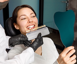 Woman in dental chair looking at reflection in mirror as dentist holds shade guide to her smile